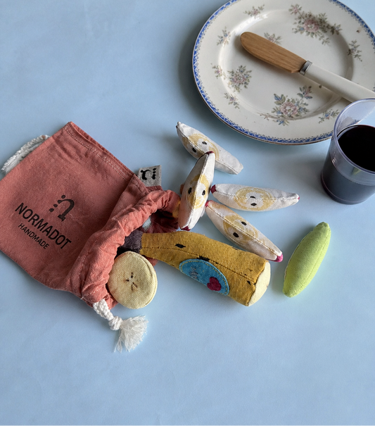 Handmade fabric play food set with a banana toy, soft play food pieces, pink Normadot drawstring bag, vintage plate, knife and glass on a light blue background.