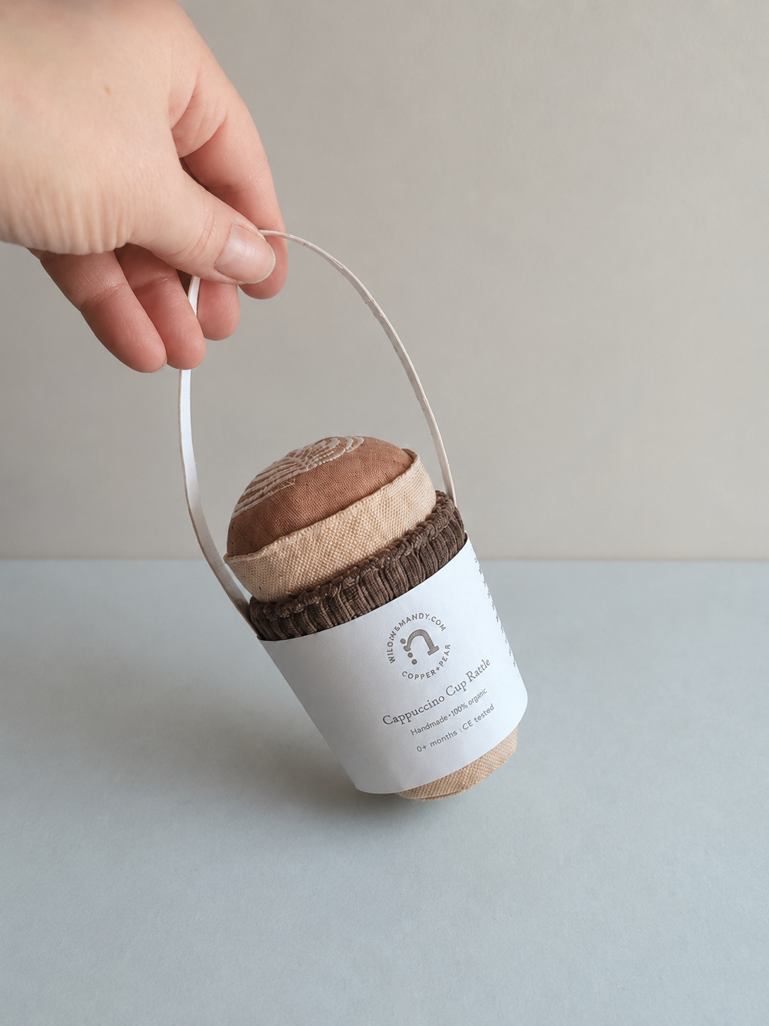 Hand holding handmade fabric play food cappuccino cup with label on neutral studio background