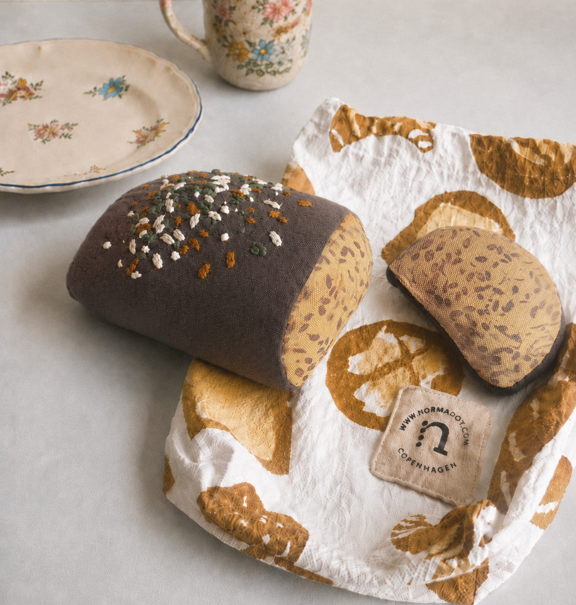 Handmade fabric rye bread loaf with one slice on a printed cotton bakery bag, styled with vintage plate and cup on a light neutral background.