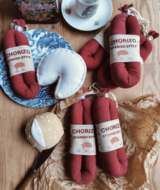 Handmade red fabric chorizo sausages with Spanish-style labels, arranged on a rustic wooden table alongside soft fabric cheeses, a blue patterned plate and crumpled brown paper.
