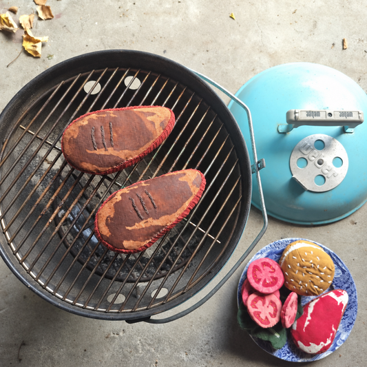 Two handmade fabric ribeye steaks with embroidered grill marks on a turquoise Weber barbecue, alongside a plate of play food including tomatoes, lettuce, burger bun and raw steak. Handmade in Denmark by normadot.