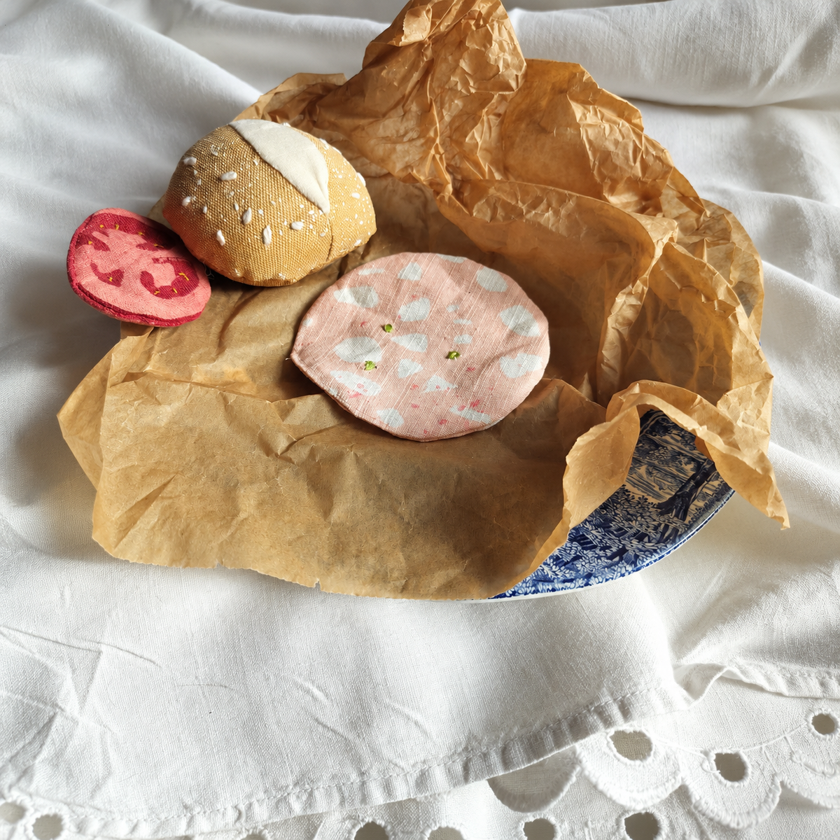 Handmade upcycled mortadella slice with embroidered pistachios, styled with a fabric bread roll and deli slice on brown paper and vintage blue-and-white plate. Textile play food by normadot.