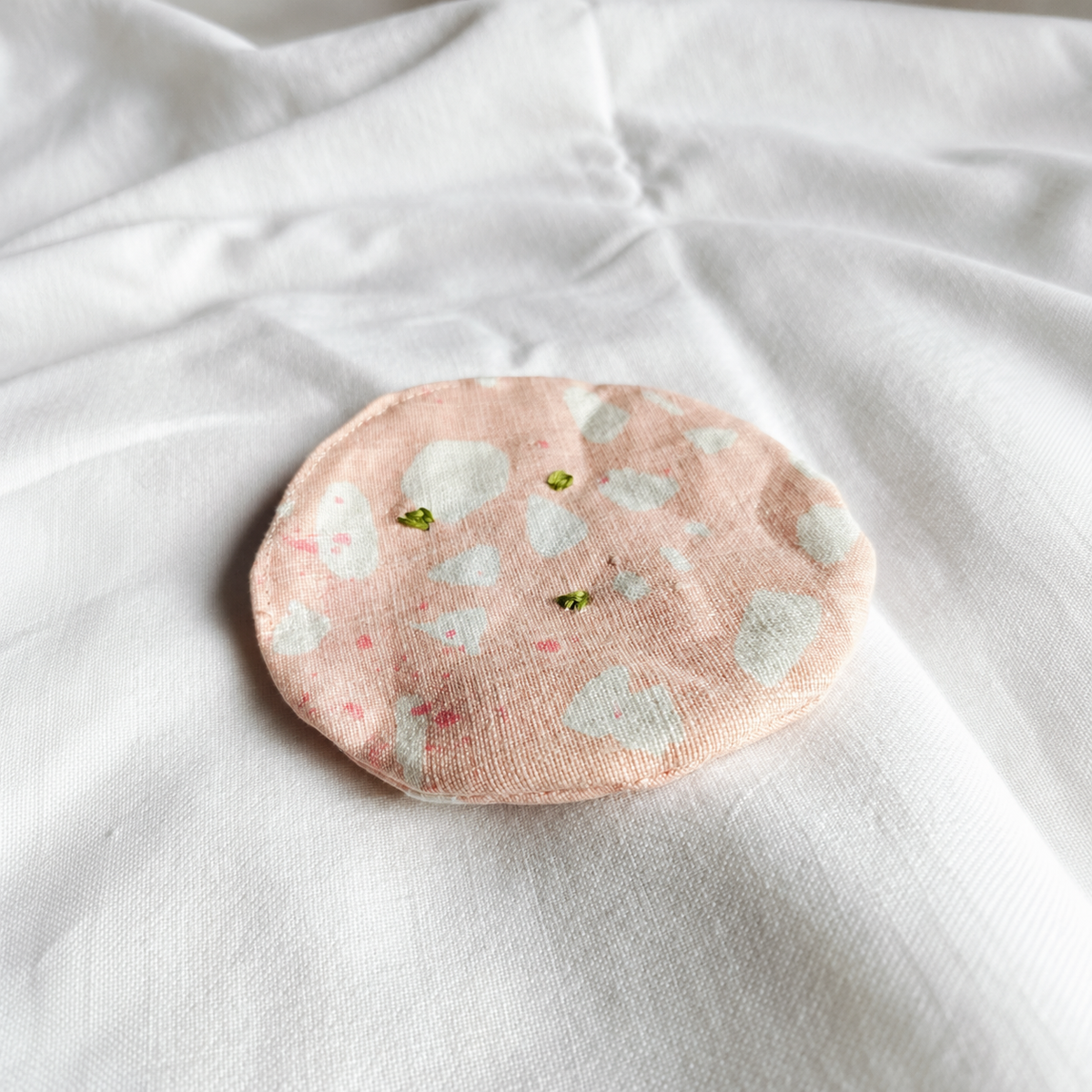 Handmade upcycled fabric mortadella slice in soft pink with white marbling and hand-embroidered green pistachios, photographed on white linen. Textile play food by normadot, made in Denmark.