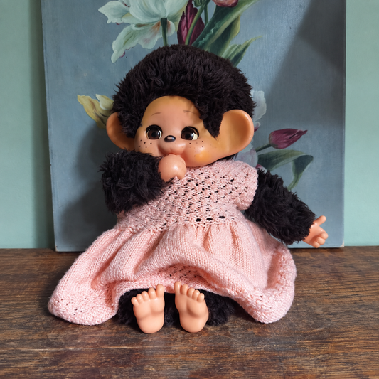 Vintage Monchhichi doll wearing a pink knitted dress, seated on a wooden table in front of a floral painting.