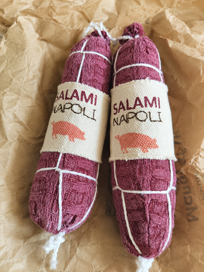 Handmade fabric Salami Napoli play food made from waffle woven cotton with printed textile labels, white thread details and recycled polyester filling, shown close-up on crinkled brown deli paper.