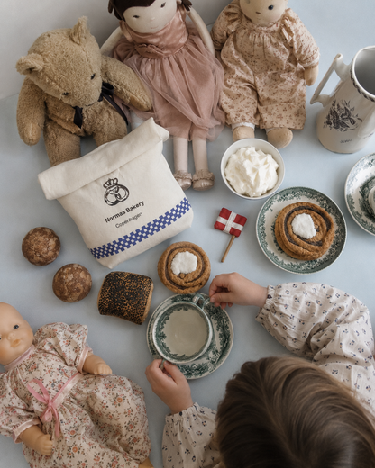Child playing with handmade Danish bakery play food, including a cinnamon roll, tebirkes and two æbleskiver, with vintage tea set, dolls and teddy bear.