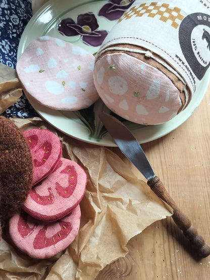 Close-up of handmade fabric mortadella and a matching slice on a floral plate, beside fabric tomato slices and brown paper on a wooden table.