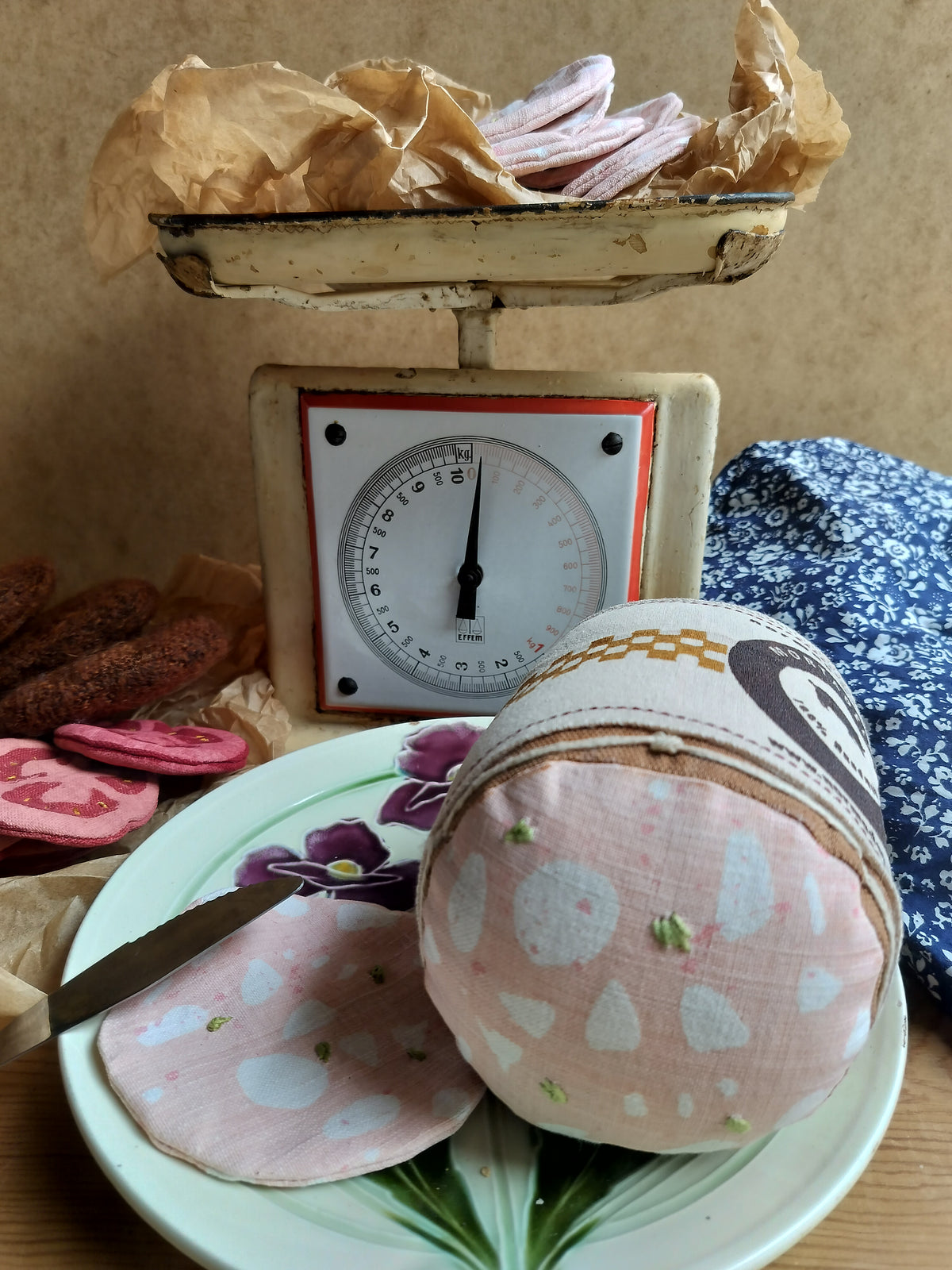 Handmade fabric mortadella and matching slice on a floral plate, styled with vintage kitchen scales, brown paper and blue floral fabric.