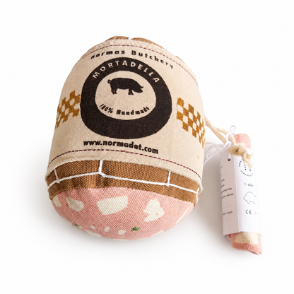 Handmade fabric mortadella with a Normas Butchery label and white string netting, beside a rolled fabric slice in a paper sleeve on a white background.